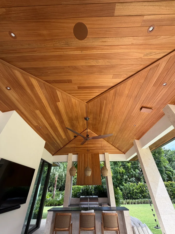 Ayous ceiling above a covered outdoor kitchen with woven pendants, stone columns and a garden view