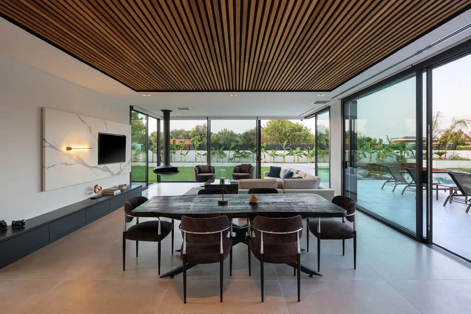 Linear wood ceiling above a dining table beside full-height garden-facing windows